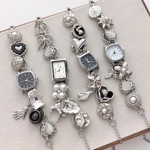 Puede incluir: Cuatro pulseras plateadas con dijes y pequeños relojes. Los relojes tienen esferas cuadradas o rectangulares en blanco o negro. Los dijes incluyen corazones, flores, lazos y perlas. Las pulseras están unidas con cadenas.