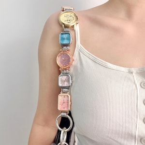 May include: A unique purse strap made of a series of colorful watches. The watches are in gold, blue, pink, and silver tones. The strap is attached to a black purse with silver hardware. The watches are connected by silver metal links.