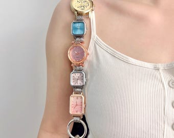 May include: A unique purse strap made of a series of colorful watches. The watches are in gold, blue, pink, and silver tones. The strap is attached to a black purse with silver hardware. The watches are connected by silver metal links.