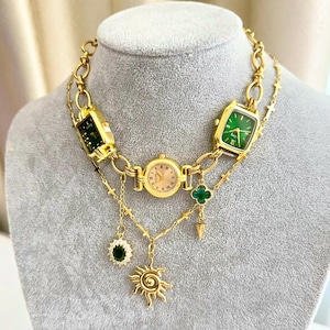 Puede incluir: Un collar dorado con tres esferas de reloj de diferentes colores. El collar tiene un diseño de cadena con colgantes, incluyendo un sol, una gema verde y un trébol de cuatro hojas.
