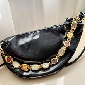 May include: A black crescent-shaped shoulder bag with a glossy finish, featuring a decorative gold-tone chain strap composed of multiple small vintage-style wristwatches in various shapes, including heart, square, oval, and hexagonal designs.