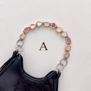 May include: Black patent leather handbag with a unique handle composed of heart-shaped watch faces in various pastel colors. The letter "A" is printed on the background.