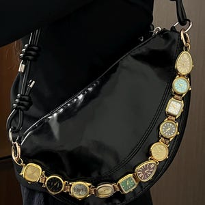 Op de afbeelding: Zwarte, halvemaanvormige handtas met een glanzende afwerking. De riem is versierd met een reeks vintage gouden horloges. De horloges hebben verschillende wijzerplaatontwerpen en kleuren, wat een unieke touch aan het accessoire geeft.