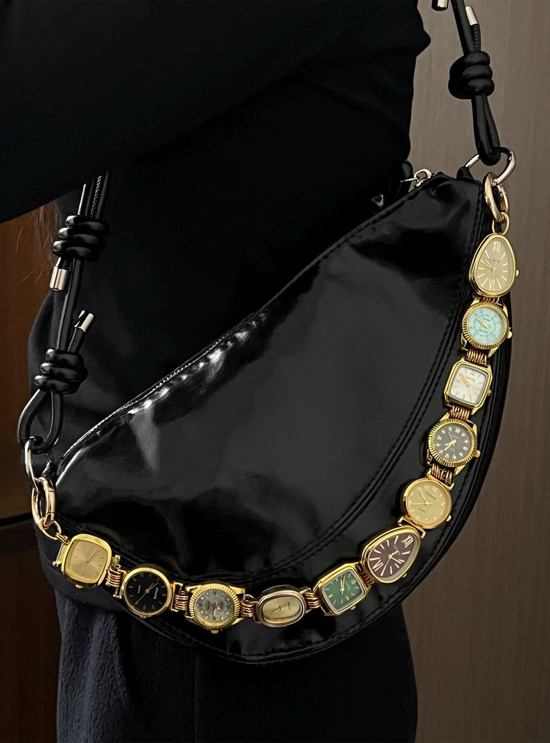 Op de afbeelding: Zwarte, halvemaanvormige handtas met een glanzende afwerking. De riem is versierd met een reeks vintage gouden horloges. De horloges hebben verschillende wijzerplaatontwerpen en kleuren, wat een unieke touch aan het accessoire geeft.