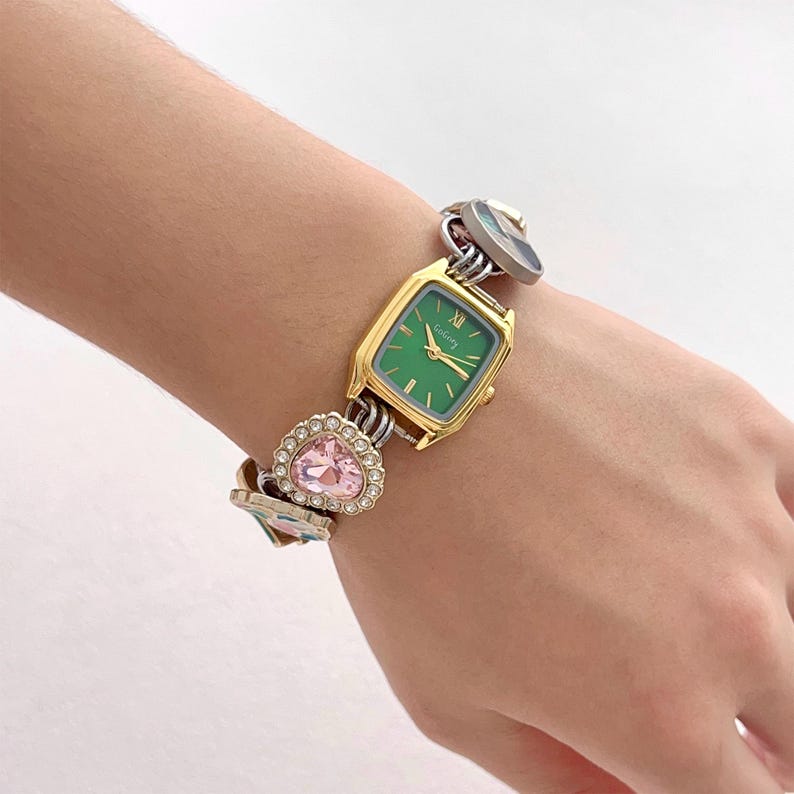 May include: A gold-toned watch with a green face and Roman numerals. The watch is part of a bracelet with heart-shaped pink crystals and other decorative elements. The watch is on a wrist.