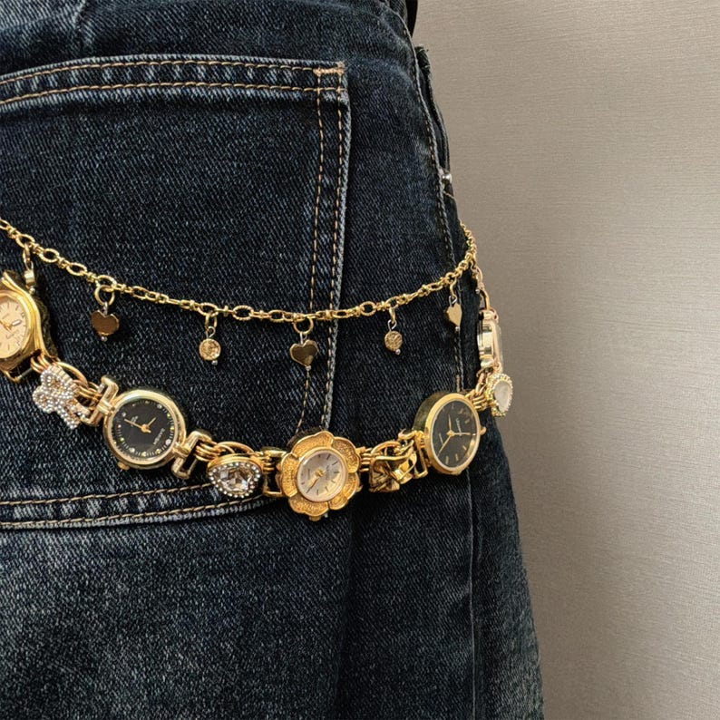 May include: A gold-toned watch belt with multiple watch faces and dangling charms, including hearts and circles, is draped over a dark denim pocket. The watches have black and gold faces, and the belt is attached to a denim garment.
