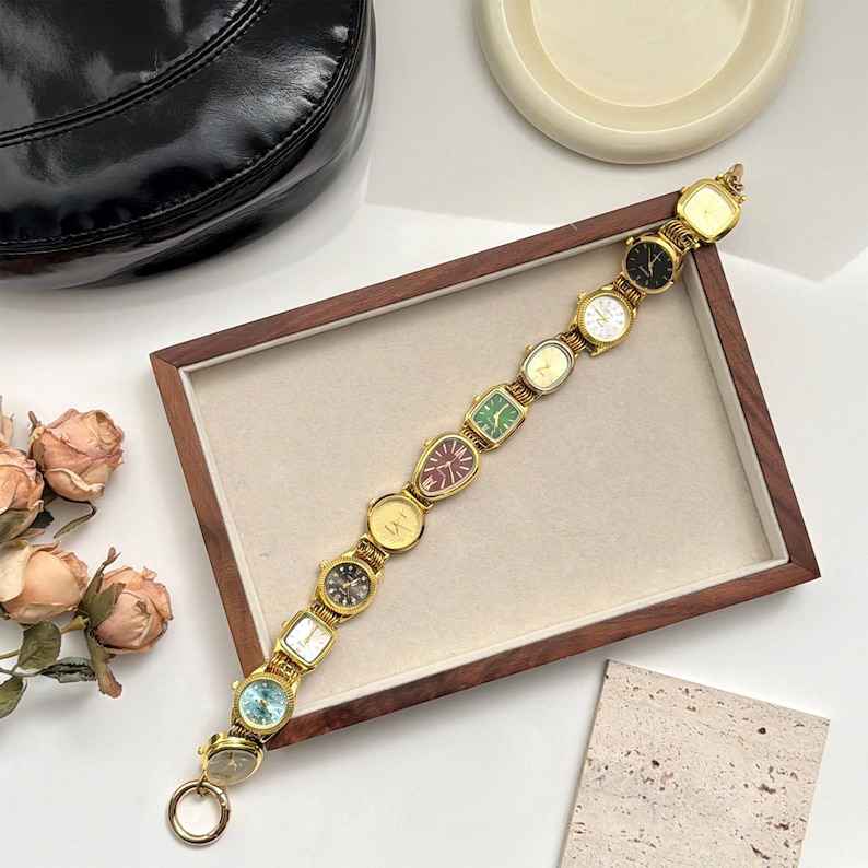 Op de afbeelding: Een goudkleurige armband met een reeks kleine wijzerplaten in verschillende vormen en kleuren. De wijzerplaten omvatten vierkante, ovale en ronde ontwerpen. De armband wordt gepresenteerd op een houten dienblad met een neutrale achtergrond.