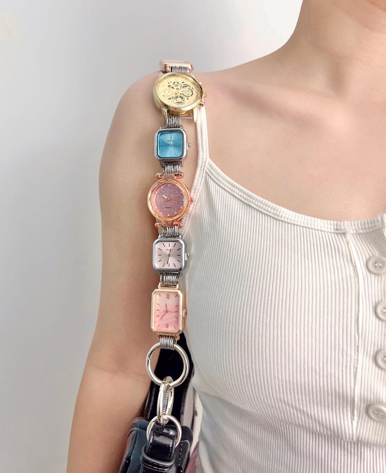 May include: A unique purse strap made of a series of colorful watches. The watches are in gold, blue, pink, and silver tones. The strap is attached to a black purse with silver hardware. The watches are connected by silver metal links.