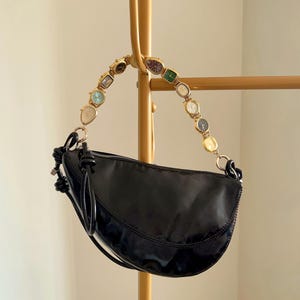 Op de afbeelding: Zwarte, halvemaanvormige handtas met een gouden kettinghandvat versierd met vintage horloges. De tas heeft een glanzende afwerking en een geknoopt detail aan de zijkant. Hij hangt aan een goudkleurige kapstok.