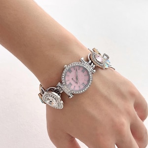 May include: A silver-toned watch with a pink face and crystal accents. The watch features Roman numerals and is connected to a bracelet adorned with decorative crystals and charms. The watch is approximately 1.25 inches in diameter.
