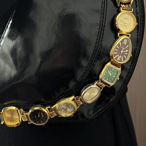 Op de afbeelding: Een zwarte handtas met een gouden ketting gemaakt van verschillende vintage horloges. De horloges hebben verschillende gekleurde wijzerplaten, waaronder goud, groen en bordeauxrood. De horloges zijn verbonden door gouden schakels.