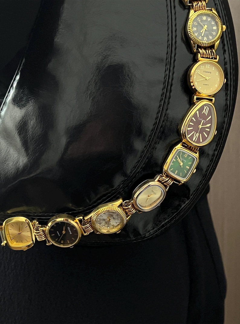 Op de afbeelding: Een zwarte handtas met een gouden ketting gemaakt van verschillende vintage horloges. De horloges hebben verschillende gekleurde wijzerplaten, waaronder goud, groen en bordeauxrood. De horloges zijn verbonden door gouden schakels.