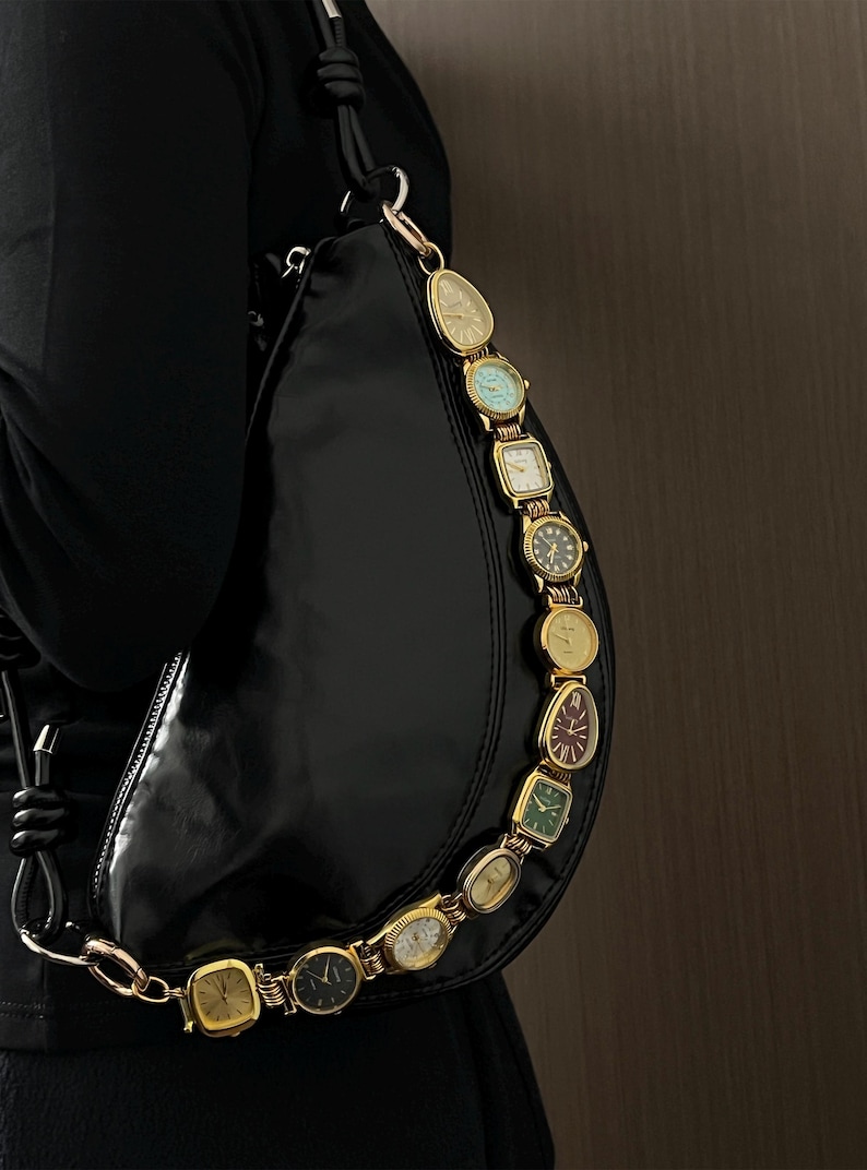 Op de afbeelding: Zwarte halvemaanvormige handtas met een gouden kettingriem bestaande uit vintage horloges. De horloges hebben verschillende vormen en kleuren, waaronder goud, groen en blauw. De tas is gemaakt van een glanzend, glad materiaal.