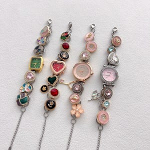 May include: Four decorative wristwatches with various embellishments. The watches feature different colored faces, including green, pink, and silver. Each watch is adorned with decorative charms, including hearts, flowers, and gemstones. The watch straps are silver chains.