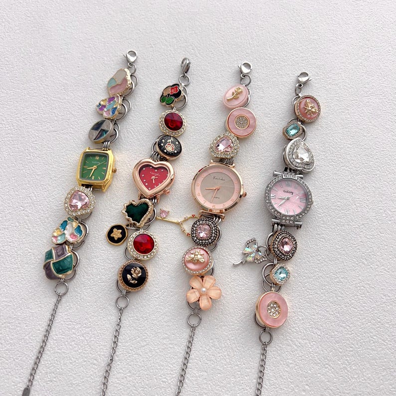 May include: Four decorative wristwatches with various embellishments. The watches feature different colored faces, including green, pink, and silver. Each watch is adorned with decorative charms, including hearts, flowers, and gemstones. The watch straps are silver chains.