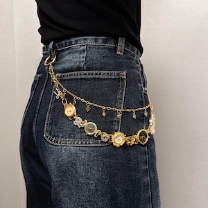 May include: Gold-toned belt featuring multiple watch faces and heart-shaped charms. The belt is attached to a pair of dark wash denim jeans. The watch faces have various designs and are connected by delicate chains.