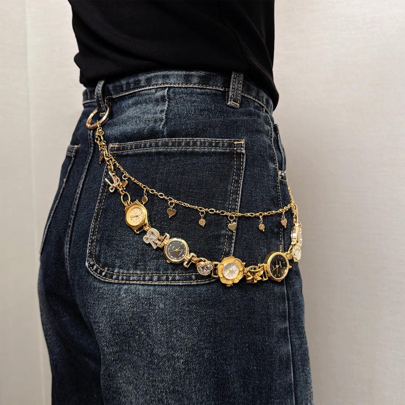May include: Gold-toned belt featuring multiple watch faces and heart-shaped charms. The belt is attached to a pair of dark wash denim jeans. The watch faces have various designs and are connected by delicate chains.