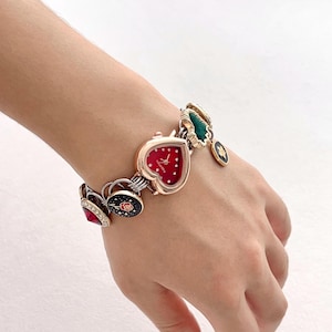 May include: A heart-shaped watch with a red face and white dots, connected to a bracelet with various charms. Charms include a rose, a green leaf, and a black and gold design. The watch and bracelet are gold and silver in color.
