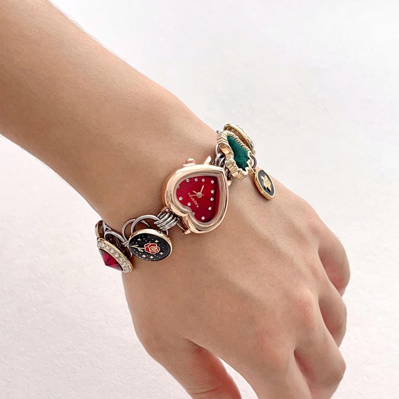 May include: A heart-shaped watch with a red face and white dots, connected to a bracelet with various charms. Charms include a rose, a green leaf, and a black and gold design. The watch and bracelet are gold and silver in color.