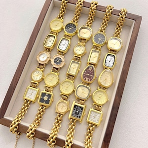 Può includere: Una collezione di orologi da polso dorati con varie forme di quadrante, tra cui rotonde, quadrate e rettangolari. Gli orologi sono esposti in un vassoio di legno, mostrando i loro intricati design e i cinturini a catena. I quadranti presentano diversi colori e stili.