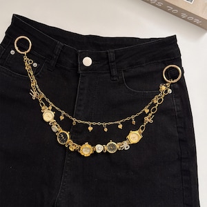 May include: A gold-colored chain belt with multiple small watch charms and heart-shaped charms. The belt is attached to a pair of black denim jeans with gold-colored rings. The watch charms have black and gold faces.