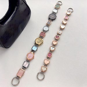 May include: Two watch straps with various watch faces, including square, rectangular, and heart-shaped designs. The straps are silver and gold-toned, with a black purse in the background. The watches have different colored faces.