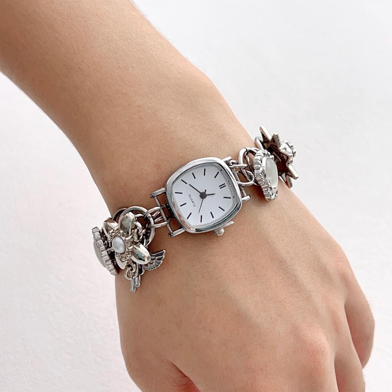 May include: Silver-toned quartz watch with a square face and white dial. The watch band features decorative floral and star-shaped charms. The watch is a stylish accessory.