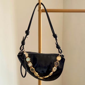 Op de afbeelding: Een zwarte, halvemaanvormige handtas met een gouden ketting van vintage horloges. De tas heeft een glanzende afwerking en zwarte leren bandjes. De tas hangt aan een goudkleurig rek.