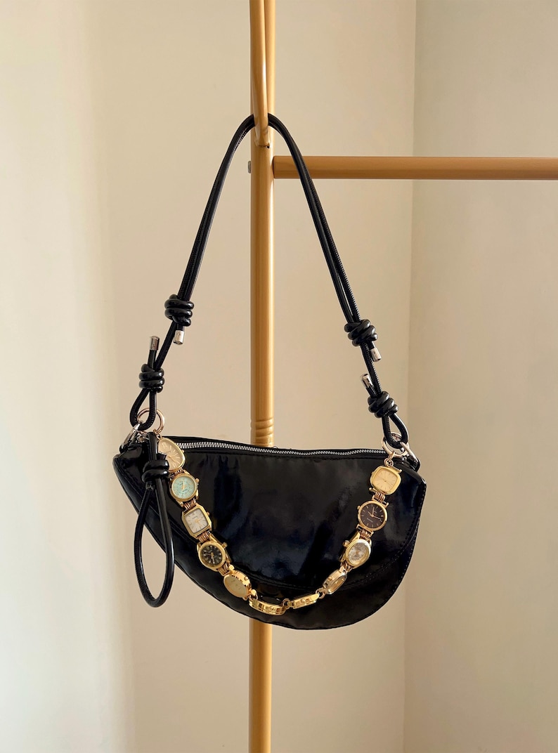 Op de afbeelding: Een zwarte, halvemaanvormige handtas met een gouden ketting van vintage horloges. De tas heeft een glanzende afwerking en zwarte leren bandjes. De tas hangt aan een goudkleurig rek.