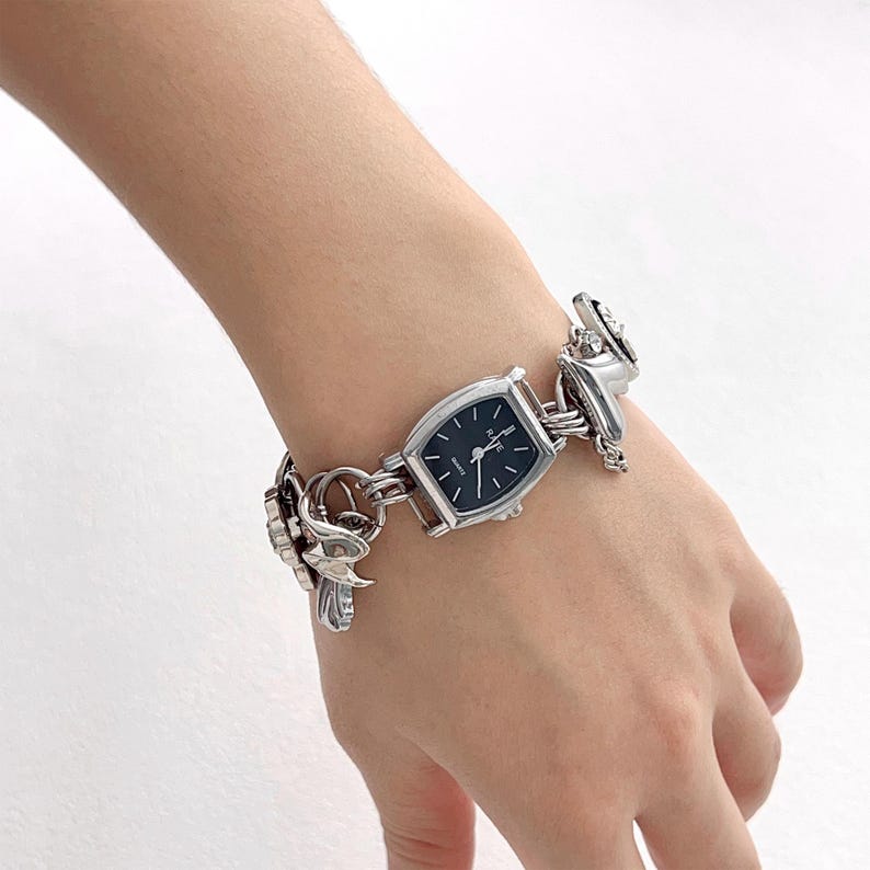 May include: Silver-toned watch with a rectangular black face and silver hands. The watch is attached to a decorative silver bracelet with ornate, stylized floral and geometric links. The watch is worn on a wrist.