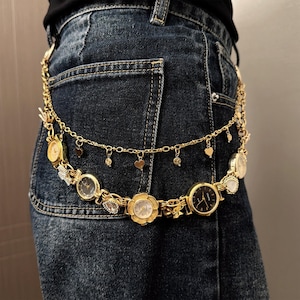 May include: A gold-toned belt featuring multiple watch faces of varying shapes and sizes. The belt includes heart-shaped charms and small dangling embellishments. The belt is worn over blue denim jeans.