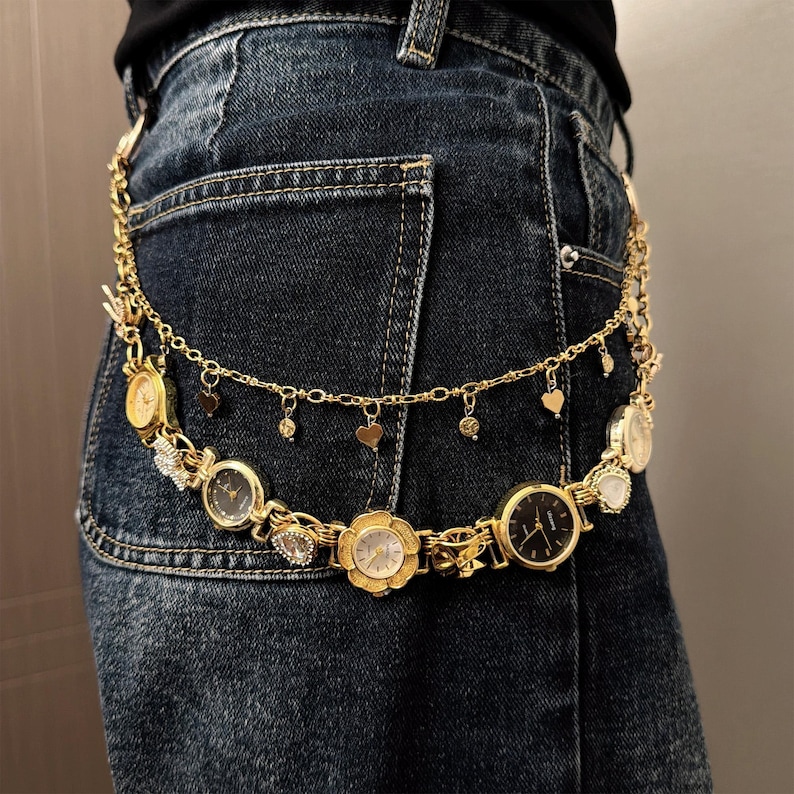 May include: A gold-toned belt featuring multiple watch faces of varying shapes and sizes. The belt includes heart-shaped charms and small dangling embellishments. The belt is worn over blue denim jeans.