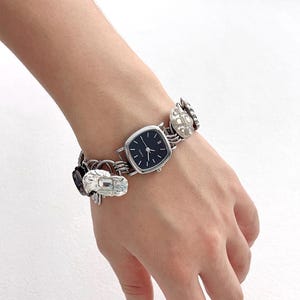 May include: A silver-toned watch with a square black face and silver hands. The watch band features decorative silver links and charms. The watch is worn on a wrist.