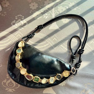 Op de afbeelding: Zwarte, halvemaanvormige handtas met een decoratieve ketting van vintage horloges. De tas heeft een zwarte leren band en een goudkleurige ketting. De horloges hebben verschillende kleuren en vormen.