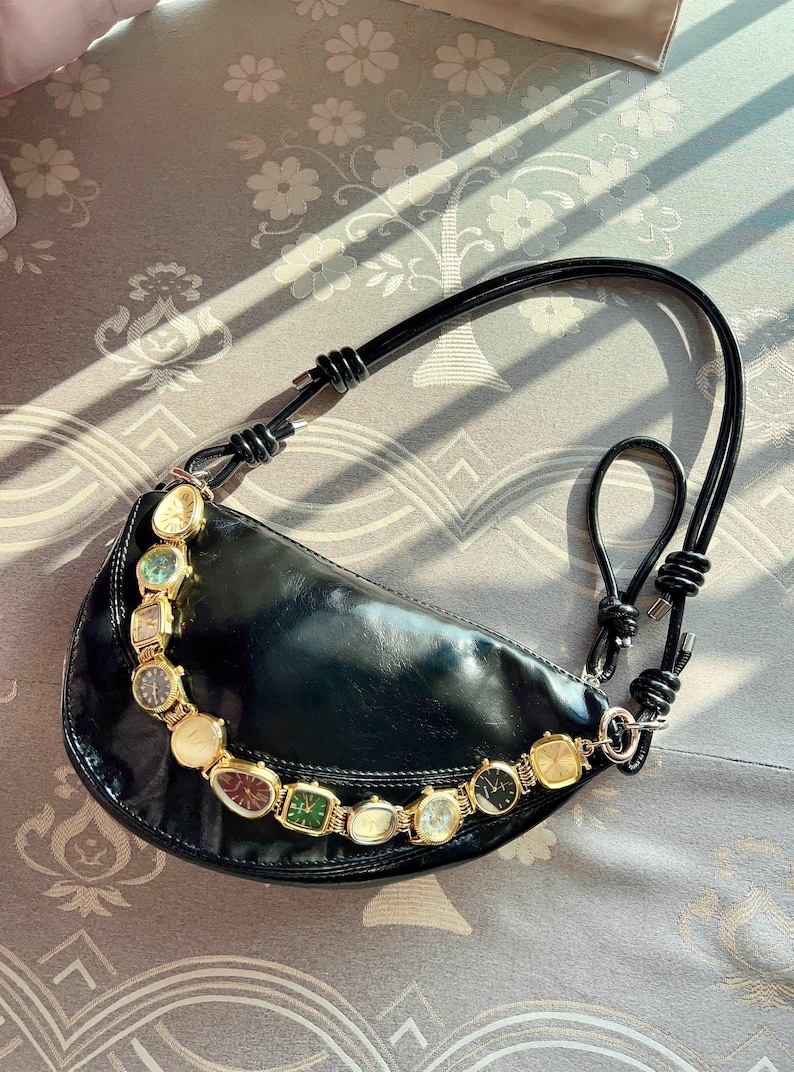 Op de afbeelding: Zwarte, halvemaanvormige handtas met een decoratieve ketting van vintage horloges. De tas heeft een zwarte leren band en een goudkleurige ketting. De horloges hebben verschillende kleuren en vormen.