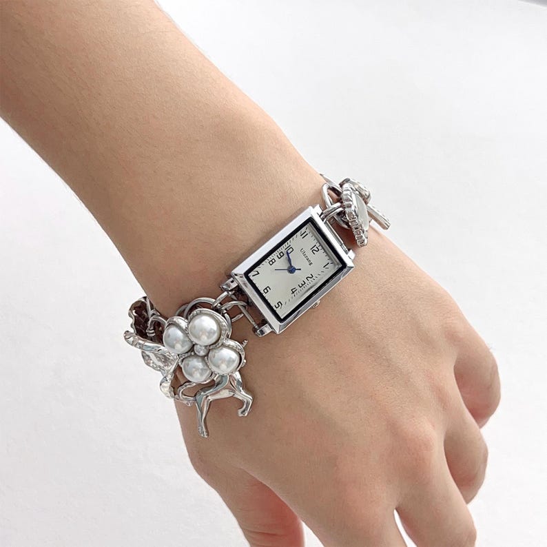 May include: Silver-toned watch with a rectangular face and a white dial. The watch band features a floral design with faux pearls and a chain-link style. The watch is worn on a wrist.