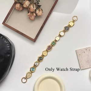 Op de afbeelding: Een goudkleurige horlogeband met meerdere kleine wijzerplaten in verschillende kleuren. De band heeft aan elk uiteinde een ronde sluiting. De tekst "Only Watch Strap" is zichtbaar.