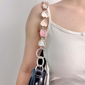 May include: A black handbag with a unique strap made of heart-shaped and rectangular watches in various pastel colors. The watches are connected by a rose gold chain and silver rings.
