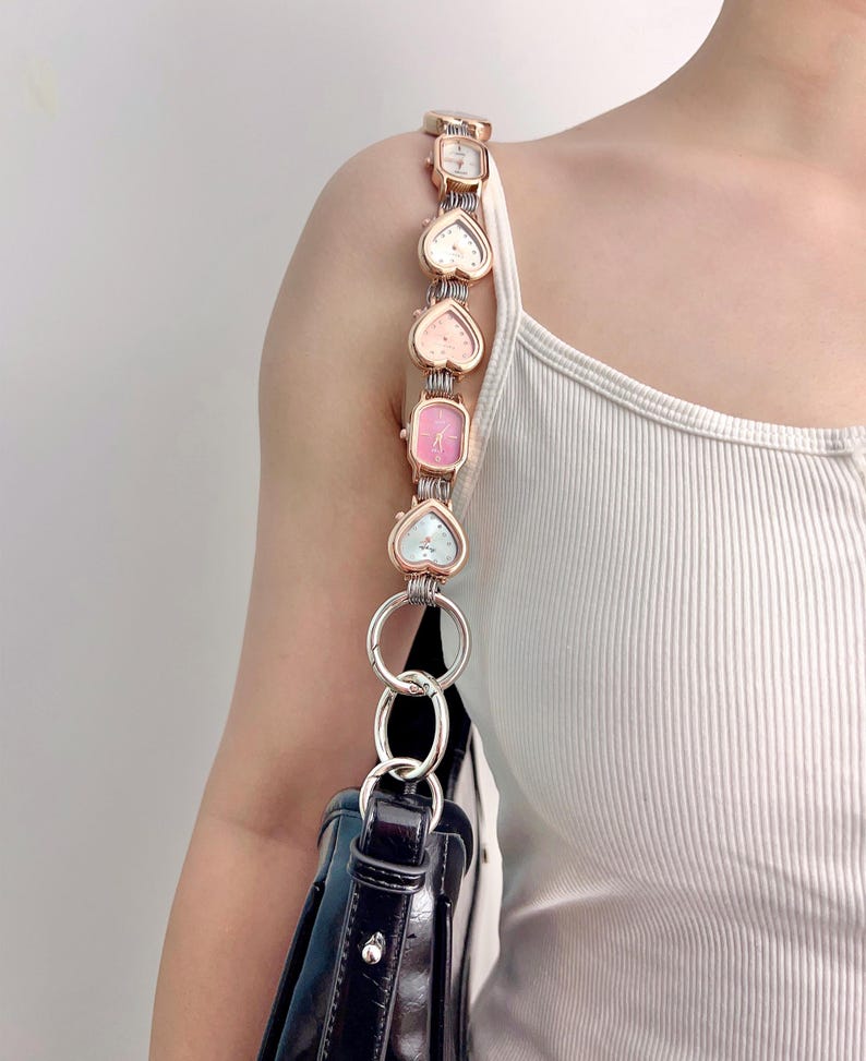 May include: A black handbag with a unique strap made of heart-shaped and rectangular watches in various pastel colors. The watches are connected by a rose gold chain and silver rings.