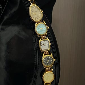 Op de afbeelding: Een zwarte handtas met een gouden kettingriem bestaande uit verschillende vintage gouden horloges. De horloges hebben verschillende wijzerplaatvormen en -kleuren, waaronder cr&egrave;me, blauw en zwart. De riem is bevestigd aan een gouden ring.