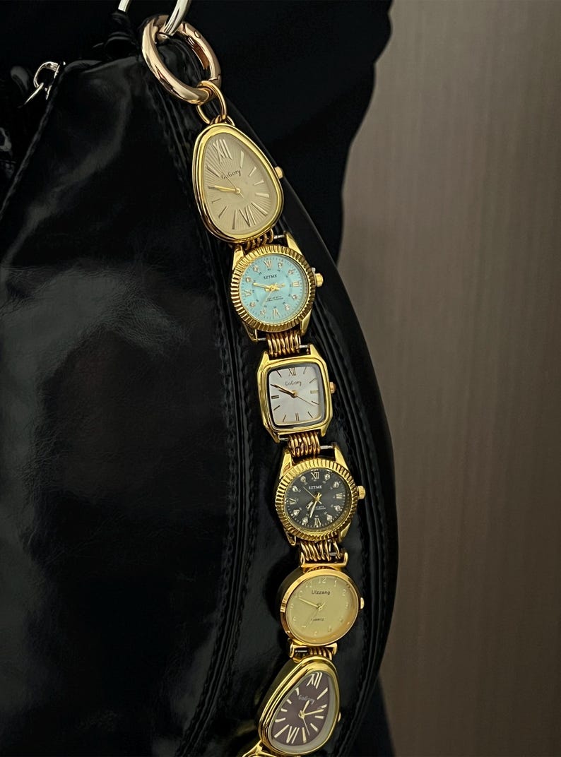 Op de afbeelding: Een zwarte handtas met een gouden kettingriem bestaande uit verschillende vintage gouden horloges. De horloges hebben verschillende wijzerplaatvormen en -kleuren, waaronder cr&egrave;me, blauw en zwart. De riem is bevestigd aan een gouden ring.
