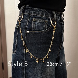 May include: Gold-colored chain belt with heart and round charms, attached to denim jeans. The chain is 15 inches long and is labeled "Style B".