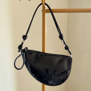 Op de afbeelding: Zwarte, halvemaanvormige handtas met een glanzende afwerking. De tas heeft een enkele handgreep en een ritssluiting. De tekst "Only Bag" staat linksboven. De tas hangt aan een goudkleurige metalen stang.