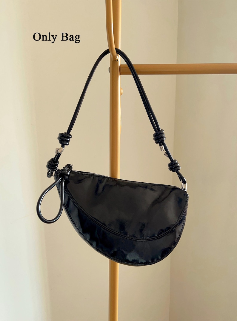 Op de afbeelding: Zwarte, halvemaanvormige handtas met een glanzende afwerking. De tas heeft een enkele handgreep en een ritssluiting. De tekst "Only Bag" staat linksboven. De tas hangt aan een goudkleurige metalen stang.