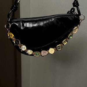 Op de afbeelding: Zwarte, glanzende, halvemaanvormige handtas met een decoratieve ketting van vintage horloges. De tas heeft een zwarte, geknoopte band en goudkleurige hardware. De horloges zijn in verschillende vormen en kleuren.