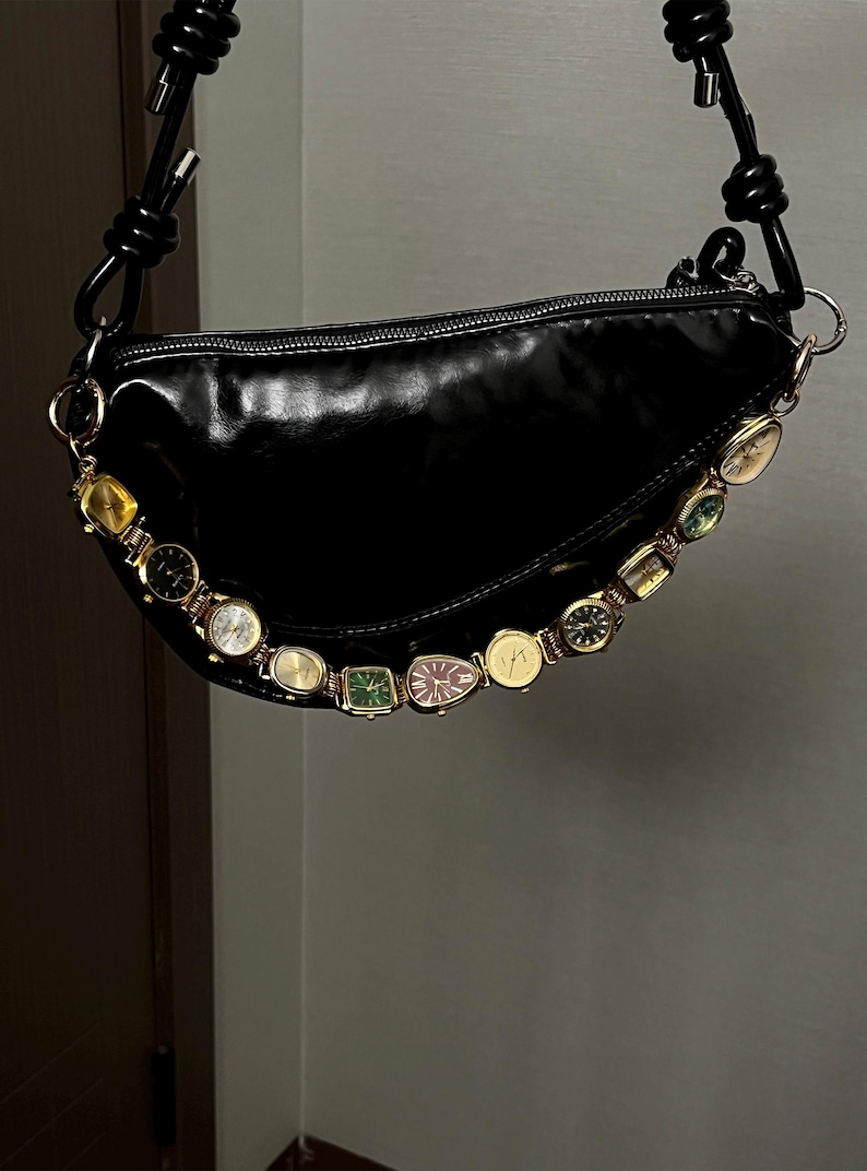 Op de afbeelding: Zwarte, glanzende, halvemaanvormige handtas met een decoratieve ketting van vintage horloges. De tas heeft een zwarte, geknoopte band en goudkleurige hardware. De horloges zijn in verschillende vormen en kleuren.