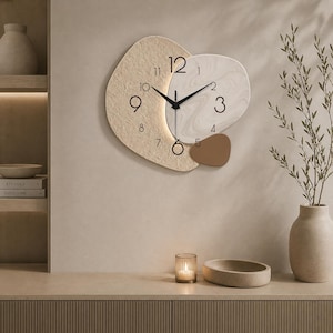 Könnte beinhalten: Abstrakte Wanduhr mit einem beige-, weiß- und braunfarbenen Design. Die Uhr hat schwarze Zeiger und Zahlen und eine einzigartige Form. Sie ist an einer neutralfarbenen Wand befestigt, mit minimalistischer Ästhetik.