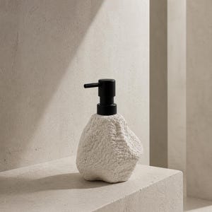 Peut inclure: Un distributeur de savon blanc texturé avec une pompe noire. Le distributeur a une forme organique, ressemblant à une pierre. La pompe est placée sur le dessus, prête à l'emploi. Le distributeur est posé sur une surface claire.