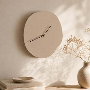 Puede incluir: Un reloj de pared beige de forma irregular con manecillas negras. El reloj está montado en una pared de color claro. El diseño del reloj es minimalista y moderno. El reloj forma parte de un tema de decoración del hogar.