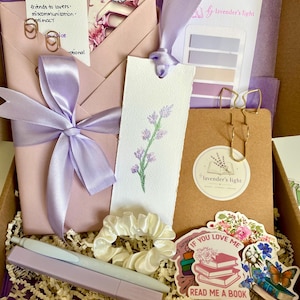 Op de afbeelding: Een cadeauverpakking gevuld met lavendel-thema items. Inclusief een roze envelop vastgebonden met een lavendel lint, een bladwijzer met een lavendel takje, stickers, een scrunchie, pennen en paperclips. De doos bevat de tekst "contemporary fiction" en "If you love me read me a book".